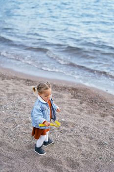 Little girl in a dress and a blue denim jacket carries sand in a toy rake on the beach near the water. High quality photo