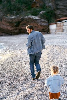 Little girl runs after smiling dad on the pebble beach against the backdrop of rocky mountains. High quality photo