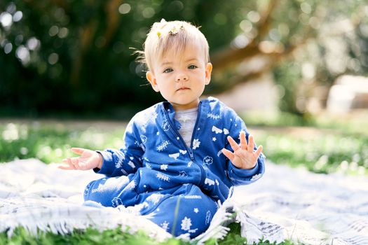 Little kid in a blue overalls sits on a blanket on a green lawn, showing his palms against a green tree background. High quality photo