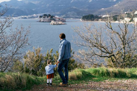 Dad holds the hand of a little girl, standing on the coast against the background of the sea, mountains and Sveti Stefan island. Back view. High quality photo