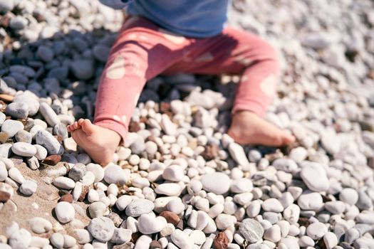 Small child in red pants is sitting on a pebble beach. Close-up. High quality photo