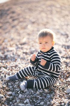 Serious toddler in striped overalls sits on a pebble beach, holding his hands in fists in front of him. High quality photo