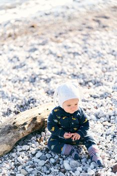 Small baby in overalls and a hat sits on a pebble beach near a tree driftwood and looks in front of him. High quality photo