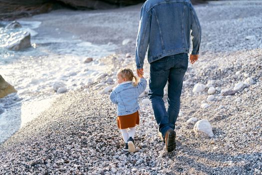 Dad leads a little daughter by the hand, walking along the pebble beach to rocky mountains and the sea. Back view. Close-up. High quality photo