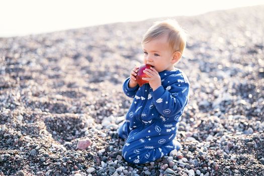 Serious kid in blue overalls sits on his knees on a pebble beach and gnaws an apple, holding it with his hands. High quality photo