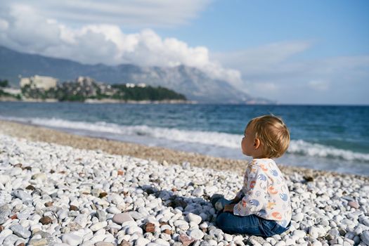Kid sits on a pebble beach by the sea. Side view. High quality photo