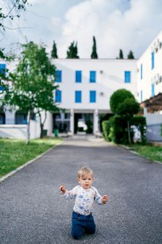 Kid is kneeling on the asphalt against the background of a building and green trees. High quality photo