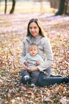 Smiling mother sits on fallen leaves and holds a baby with a rattle in her arms. High quality photo