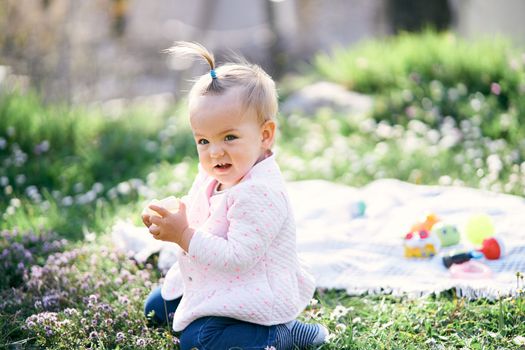 Little girl with a ponytail on her head sits on a green lawn among the wildflowers and holds a cube in her hands against the background of a bedspread with toys. High quality photo