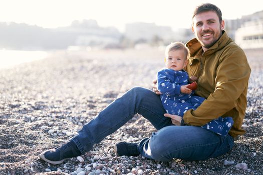 Smiling dad in a brown jacket and jeans holds a little kid in a blue overalls with an apple in his hands while sitting on a pebble beach. High quality photo