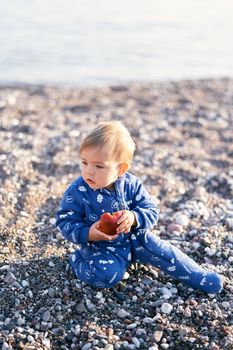 Small baby in a blue overalls sits on a pebble beach and holds an apple. Close-up. High quality photo