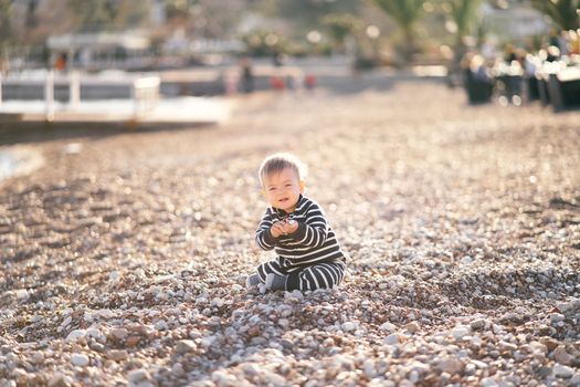 Cute toddler in striped jumpsuit sitting on a pebble beach holding his hands in front of him. High quality photo