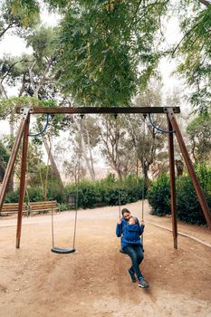 Mom in a blue warm sweater and jeans swinging on a swing with a tiny baby on her chest in the park. High quality photo