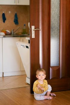 Cute kid in a striped blouse sits on the kitchen floor against the background of an open door and a dishwasher behind it. High quality photo