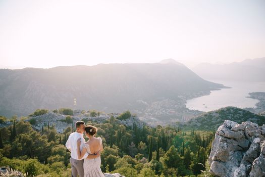 Groom hugs the bride on a high mountain against the backdrop of a panorama of the bay and trees. High quality photo