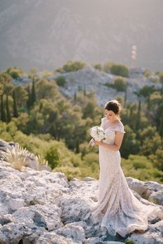 Bride with a bouquet of flowers stands on the rocks against the backdrop of mountains and greenery. High quality photo