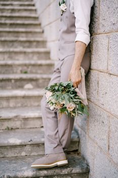 Groom with a bouquet stands on the stone steps, leaning against the wall. High quality photo