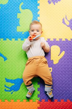 Tiny baby in a white bodysuit and brown pants lies on a colored rug, holds and gnaws on a bright rattle. Top view. High quality photo