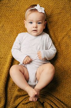 Tiny baby girl with a flower on her head in a white bodysuit lies on a yellow blanket with her hands on her tummy. High quality photo