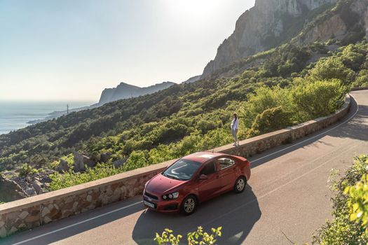 Mountain road woman. She walks along a parapet road in the middle of a mountainous area, a car is parked nearby. She is dressed in jeans and a white shirt, her hair is braided