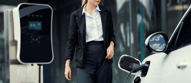 Closeup progressive businesswoman wearing suit with electric car recharging at public charging station at modern city center. Eco friendly rechargeable car powered by alternative clean energy.