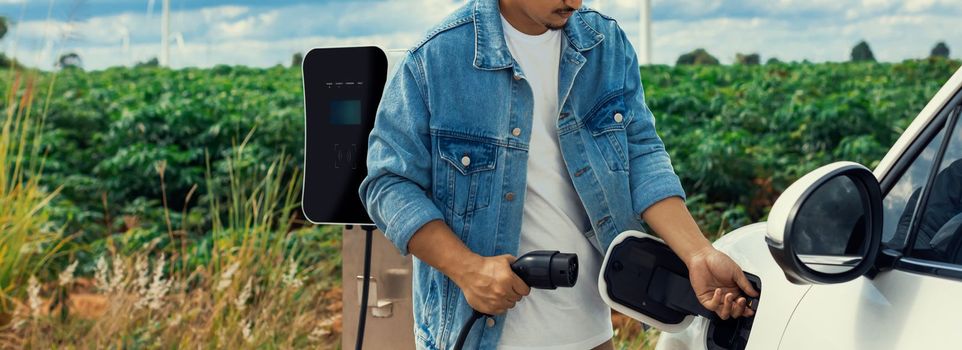 Progressive man with his electric car, EV car recharging energy from charging station on green field with wind turbine as concept of future sustainable energy. Electric vehicle with energy generator.