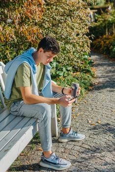 a teenager sits on a bench in the park drinks coffee from a thermo mug and looks into a phone. Portrait of handsome cheerful guy sitting on bench fresh air using device browsing media smm drinking latte urban outside outdoor.