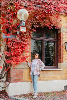 a beautiful girl stands against the background of the window of an old European house, entwined with a floating red color in autumn. Tourism & Travel Concept. Nice portrait of a young woman, in boho style outdoors in fall autumn day. Caucasian female girl 20 30 years old