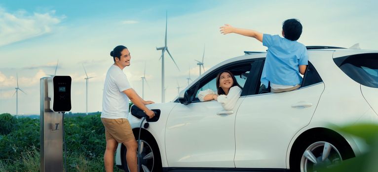 Concept of progressive happy family enjoying their time at wind farm with electric vehicle. Electric vehicle driven by clean renewable energy from wind turbine generator for charging station.