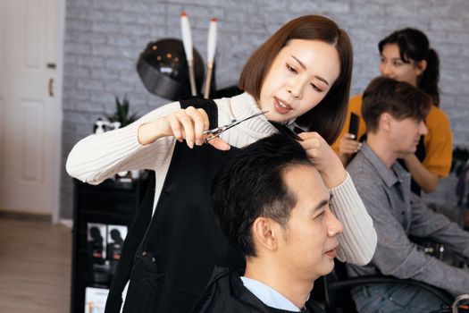 Asian female hairdresser making haircut for two male customer with male hairstyle in qualified barbershop. Men's hairstyling by a scissor and comb in hair salon concept.