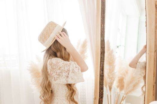 A beautiful teenage girl with long hair measures a straw hat in front of a mirror. Self-admiration of a blonde. selective focus