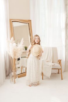 A beautiful teenage girl with long hair measures a straw hat in front of a mirror. Self-admiration of a blonde. selective focus