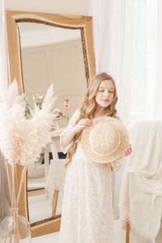 A beautiful teenage girl with long hair measures a straw hat in front of a mirror. Self-admiration of a blonde. selective focus