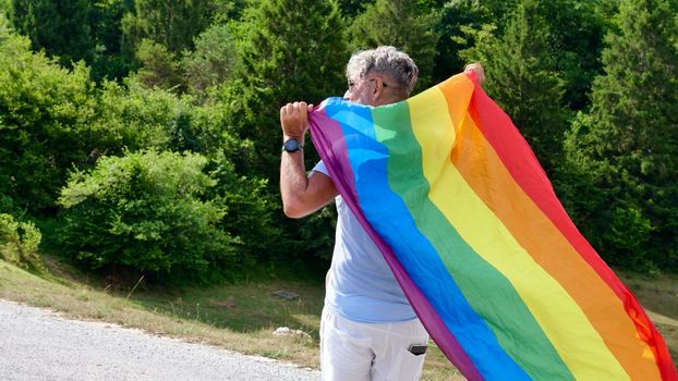 Bisexual, gay, old man, male, transsexual walk back with LGBTQIA flag, rainbow peace in pride mounts on the nature on a day and celebrate Bisexuality Day or National Coming Out Day