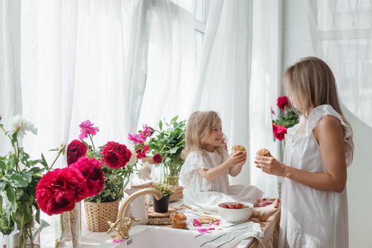 A little blonde girl with her mom on a kitchen countertop decorated with peonies. The concept of the relationship between mother and daughter. Spring atmosphere.