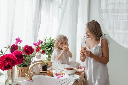 A little blonde girl with her mom on a kitchen countertop decorated with peonies. The concept of the relationship between mother and daughter. Spring atmosphere.