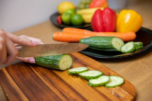 Autumn colored vegetables cut by a woman on a brown board. She slices peppers and carrots with a sharp knife.