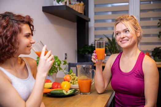 Two friends sitting in the kitchen at the table. The women in the meeting are holding glasses with orange juice. in the background a curtained window, on the table herbs in a pot.