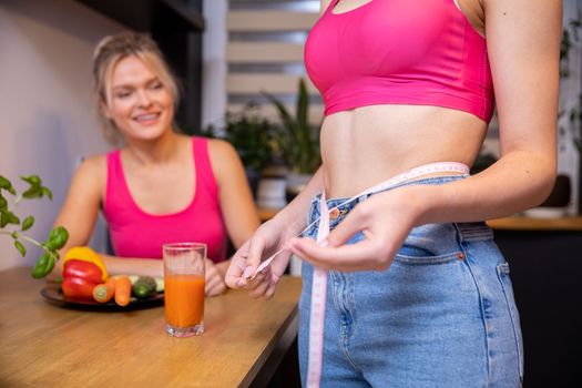 A blurry female figure in the background looks at the flat belly of her friend standing in profile. A woman's torso is visible in the foreground measuring the circumference of her slim waist.