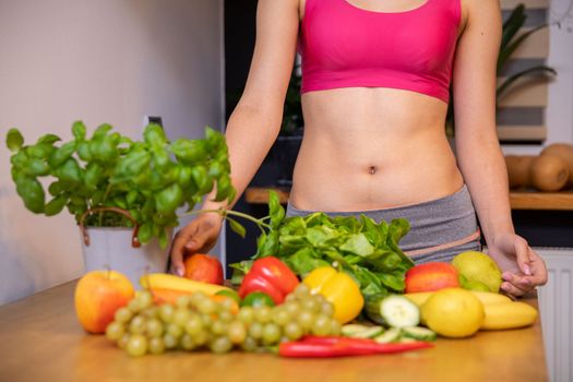 An athletic woman stands behind a table on which vegetables, fruits and herbs lie. She is dressed in a short sports tight top and only her torso is visible. Her muscular belly is exposed.
