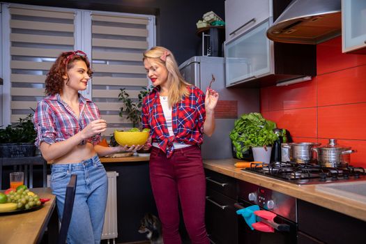 Two young women preparing a meal in the kitchen. The brunette shows the blonde the contents of the yellow bowl she is holding. Both women are wearing checkered shirts. A small dog stands on the floor.