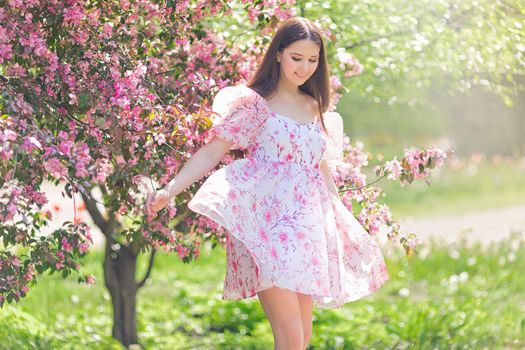 Pretty brunette girl in light pink dress, dancing near a pink blooming apple trees, in the summer in the garden in sanny day. Copy space