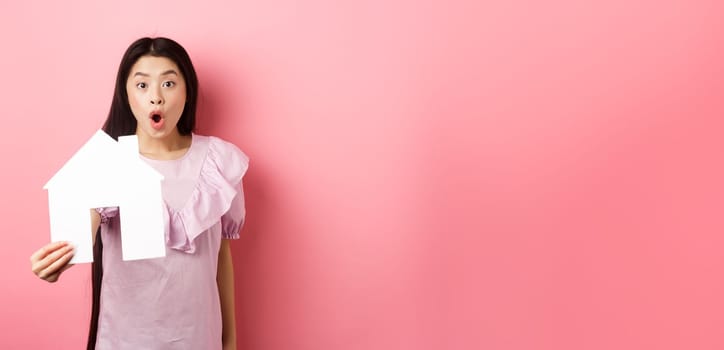 Real estate and insurance concept. Excited asian woman showing paper house cutout and look amazed, special offer, standing on pink background.