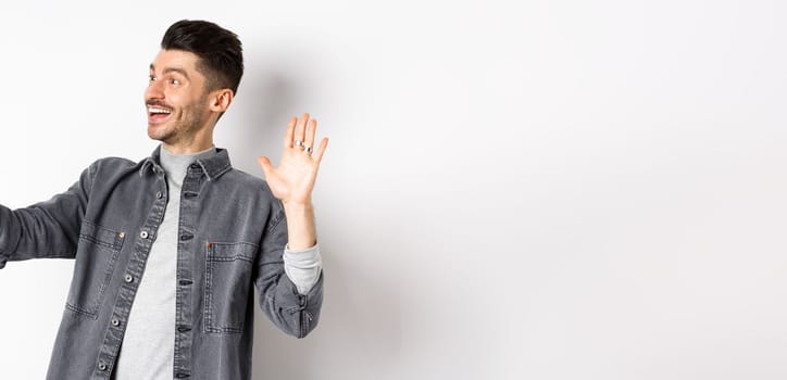 Young man video chatting on mobile phone, waiving hand at smartphone camera, greeting friend online, standing on white background.
