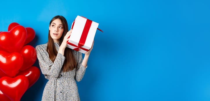 Valentines day. Beautiful woman shaking gift box to guess what inside, look dreamy, celebrating lovers holiday, standing near red hearts, blue background.