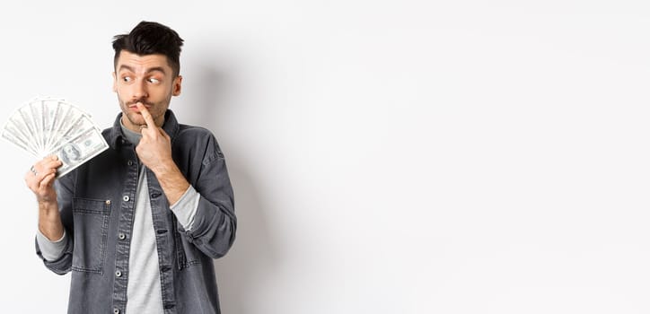 Man thinking of shopping while looking at dollar bills, holding money and staring pensive, standing on white background.