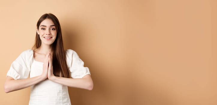 Pretty please. Cute young woman asking for favour, holding hands in begging pose and smiling, need help, standing on beige background.