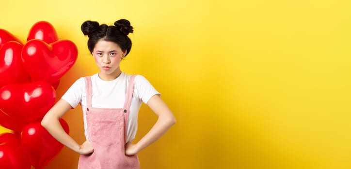 Valentines day and relationship concept. Disappointed asian girlfriend sulking, looking offended at camera, mad at boyfriend, standing near heart balloons, yellow background.