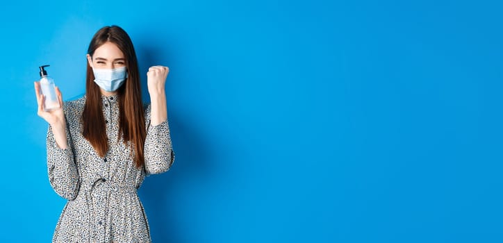 Covid-19, social distancing and healthcare concept. Motivated and excited girl in medical mask cheering, showing bottle of hand sanitizer and say yes, standing on blue background.