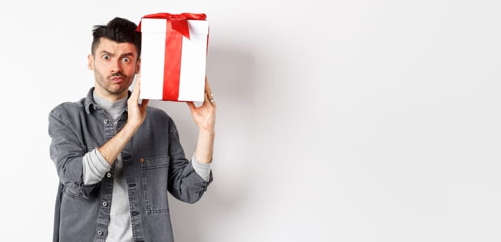 Intrigued young man looking confused at camera, trying to guess what inside gift box from lover, shaking present and stare puzzled, standing on white background. Valentines day concept.